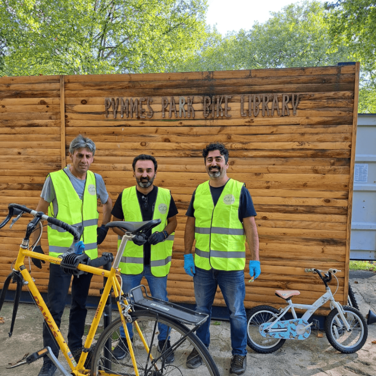 L-R Mustafa, Ergin and Ozgur standing around a bike repair in front of their shed space