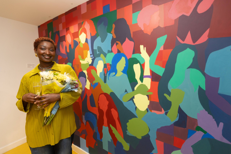 Chikaora Obiora standing next to her wall mural of lots of colourful people moving in a crowd